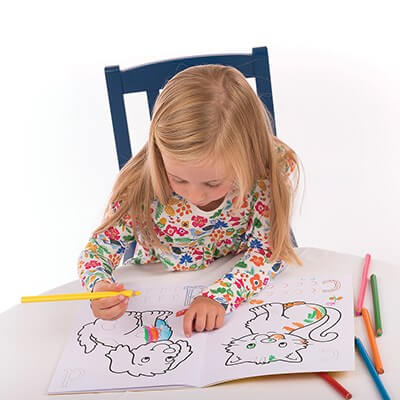 A girl in a floral shirt colors a dog and cat in an alphabet book with colored pencils on a white table.