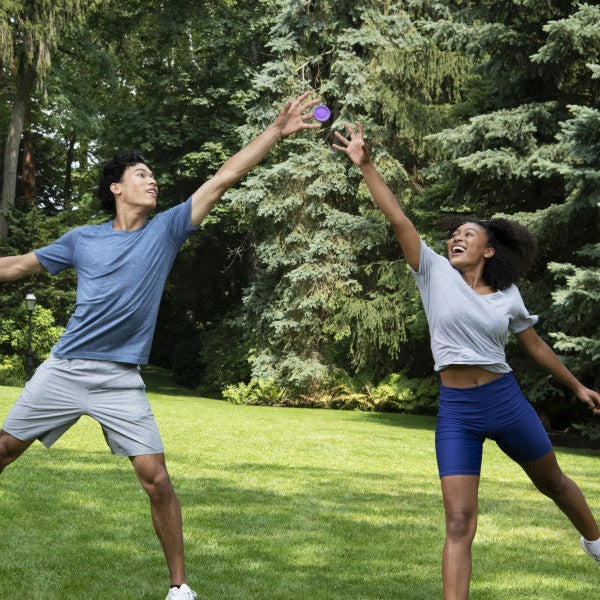 A man and woman reach up to catch a small purple circular flying disc in a grassy field with trees.