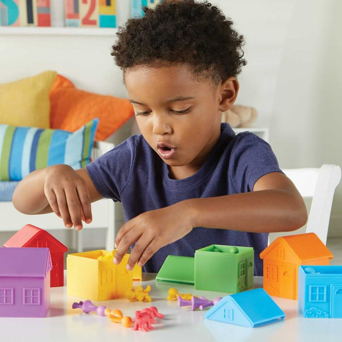 A young boy sorts colorful family figurines into matching cube-shaped toy houses on a white tabletop.