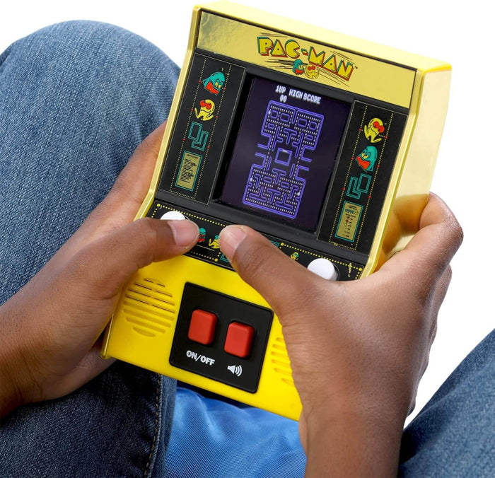 Yellow handheld Pac-Man arcade game with a color screen and red buttons, held and played by a person in blue jeans.