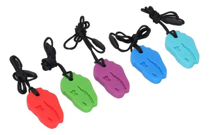 Five dinosaur-shaped silicone chew pendants in red, green, purple, blue, and light blue on black breakaway cords.