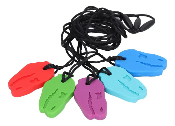 Five dinosaur head-shaped silicone chew necklaces in red, green, purple, teal, and blue on black cords.