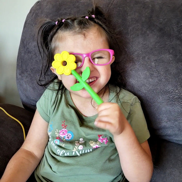 A smiling young girl holding a yellow flower chew toy with a green leafy stem up to her face.