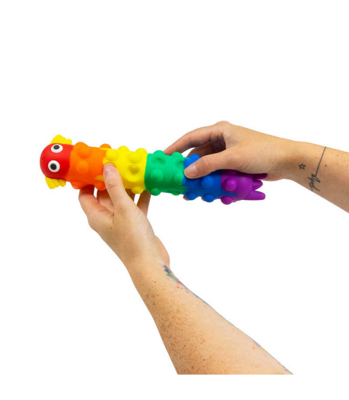 Hands hold a long, rainbow-colored segmented silicone toy with an axolotl head and suction cup bubbles.