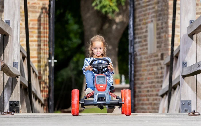 A girl rides a grey pedal go-kart with bright orange chunky tires and red shocks on a wooden walkway.