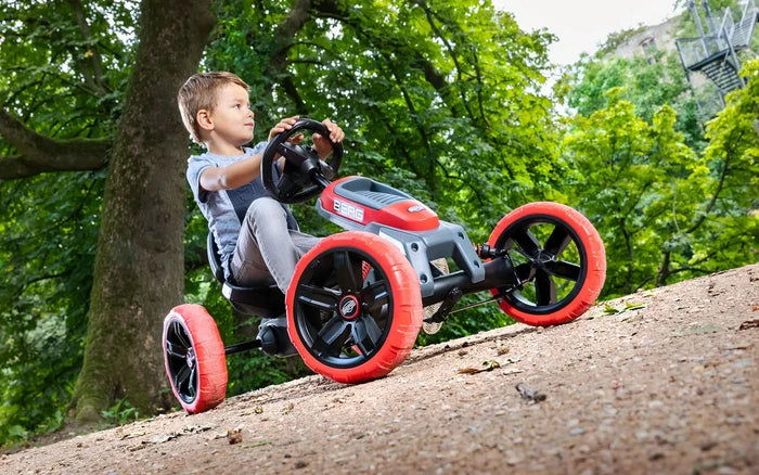 Young boy riding a red and grey BERG pedal go-kart with chunky red tires on a dirt path in a wooded park.