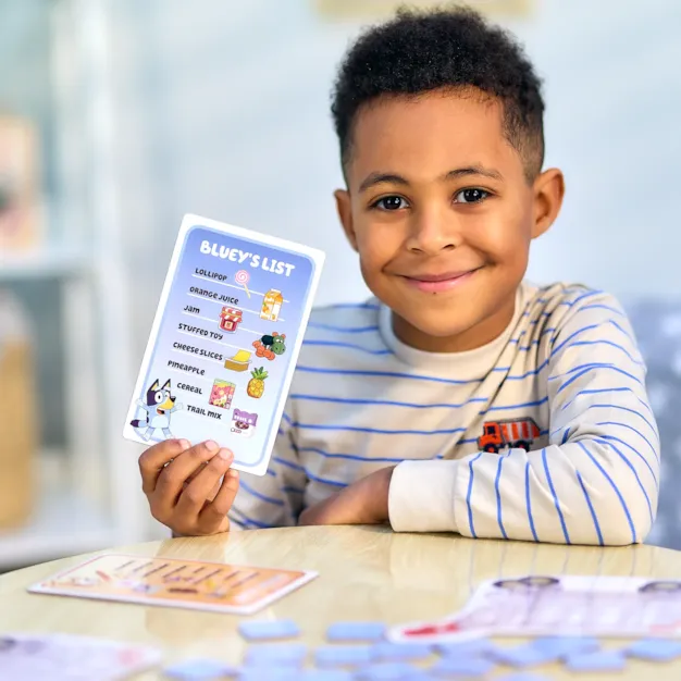 Young boy holding a Bluey shopping list card with food illustrations over a table with blue game tiles.