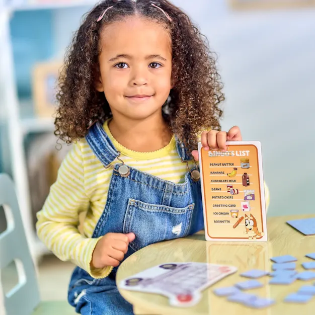 Young girl in denim overalls holding a Bluey Bingo's List card at a table with square blue game tokens.