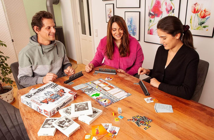 Three people playing Bomb Busters board game with cartoon rabbit art, cards, and mission boxes on a wooden table.