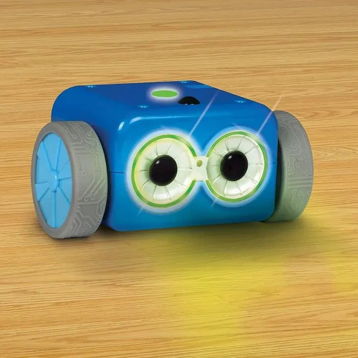 Blue cube-shaped toy robot with large glowing green eyes and grey treaded wheels on a wooden floor.