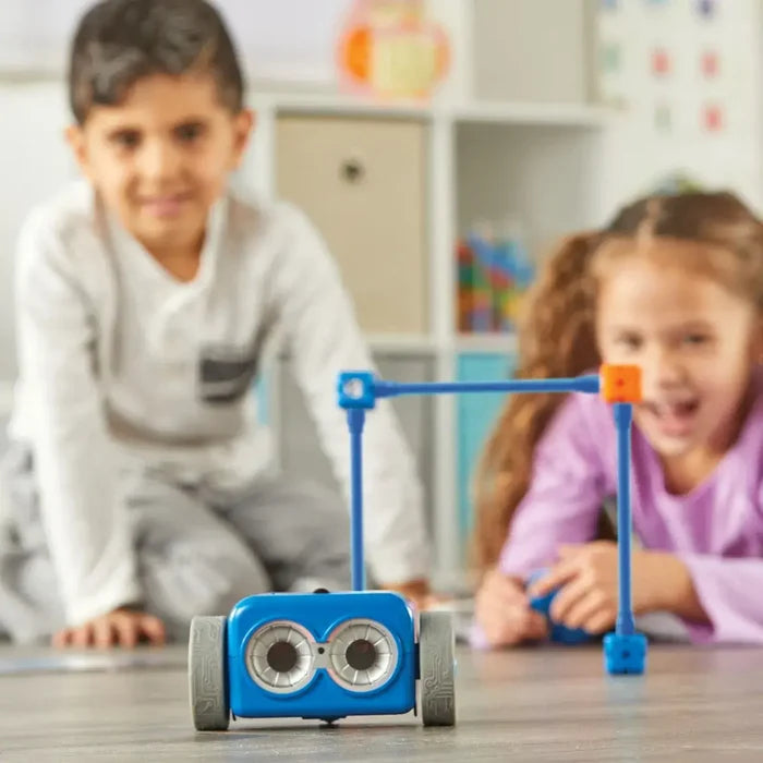 Blue rectangular toy robot with large eyes facing a plastic goal post with two children smiling in the background.