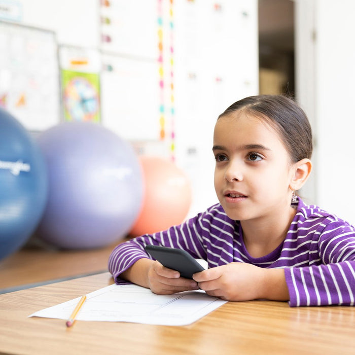 Child at a desk holding a dark gray phone-shaped rubber fidget toy with large therapy balls in the background.