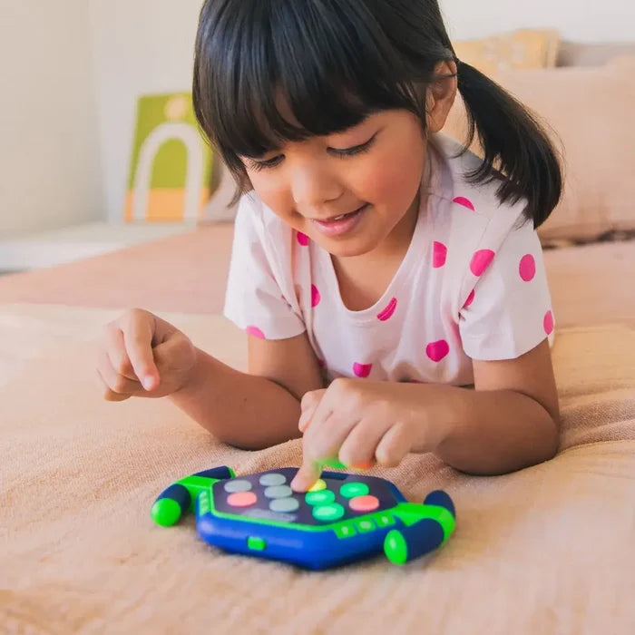 Young girl playing with a blue hexagonal handheld game featuring green side handles and glowing colorful buttons.