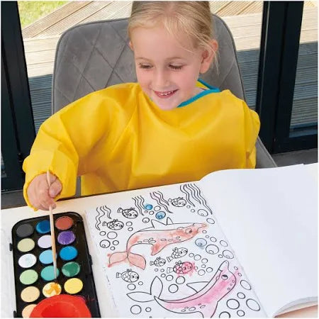 A child in a yellow smock uses watercolors to paint dolphins and fish in an open coloring book on a table.