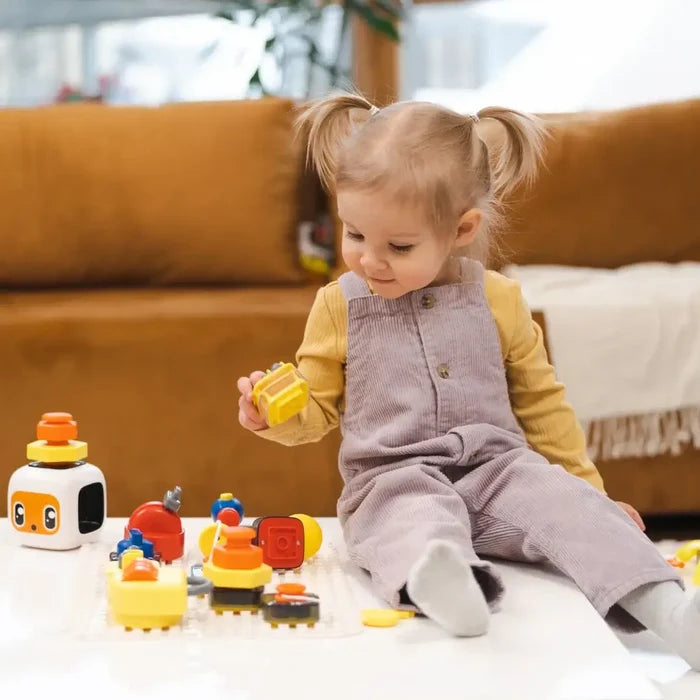 Toddler plays with a colorful toy set featuring knobs, buttons, and a white robot cube on a white table.