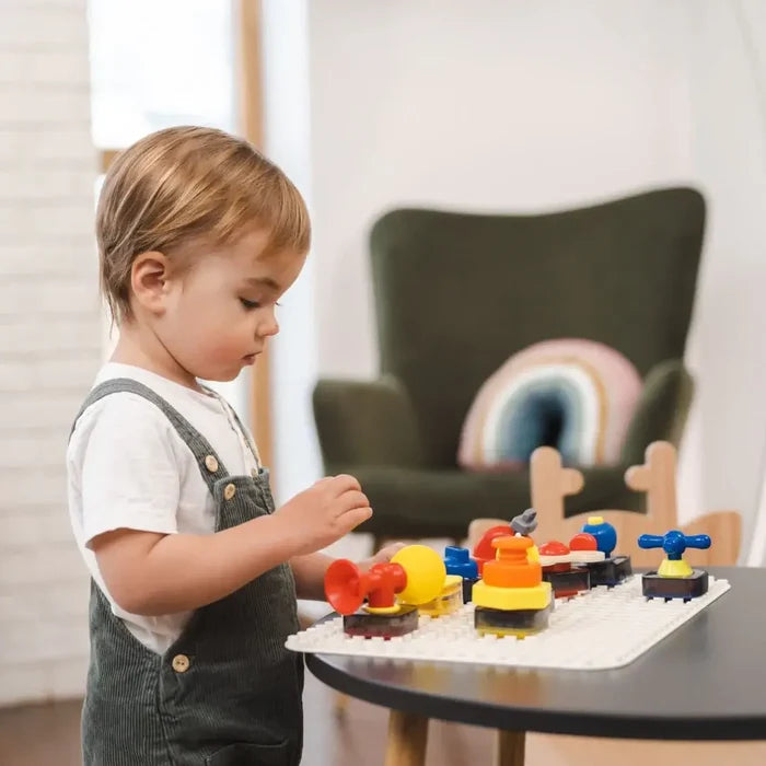 Toddler playing with a white sensory board featuring colorful red, yellow, and blue tactile knobs and buttons.