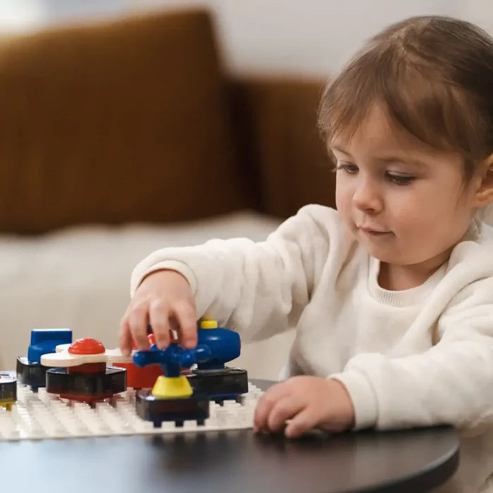 Toddler girl turns a blue cross-shaped dial on a white sensory board with colorful red and yellow knobs on a black table.