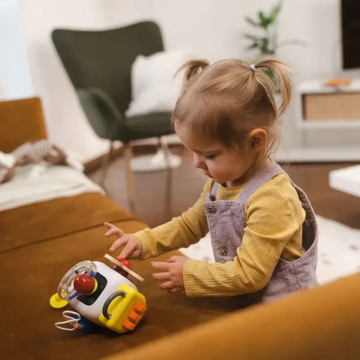 Toddler in purple overalls playing with a colorful white and yellow busy board toy featuring buttons and dials.
