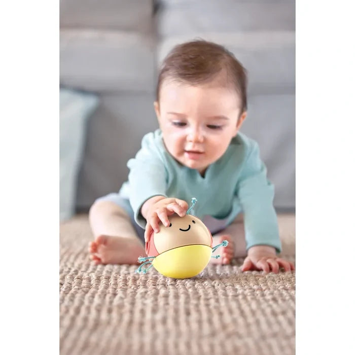 Baby playing with a round yellow and peach bug toy with a smiling face and teal cord legs on a neutral rug.