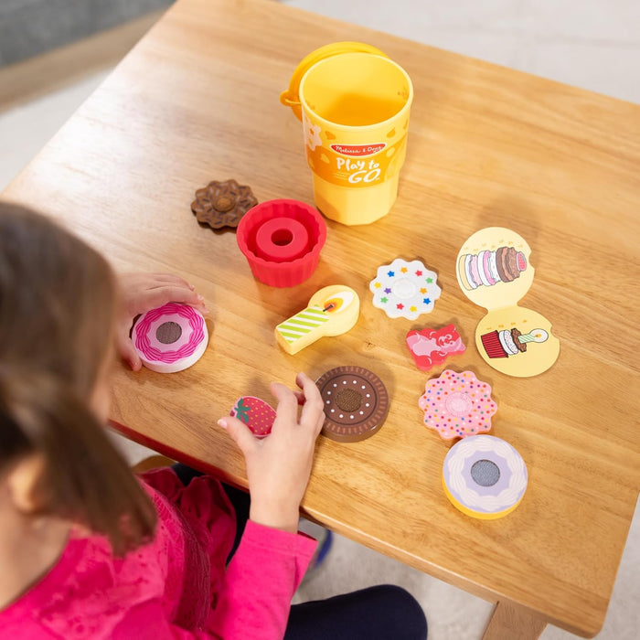 Child playing with colorful wooden toy treats, a red cupcake mold, and a yellow storage cup on a wooden table.