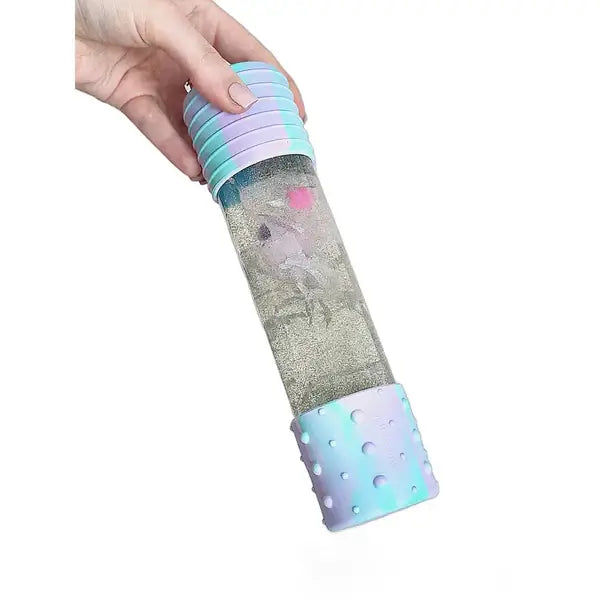 Cylindrical sensory bottle with pastel blue and purple textured caps, swirling silver glitter, and pink unicorn shapes.