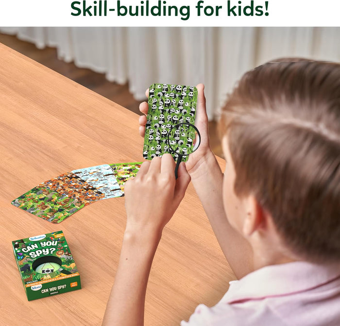 Child uses a magnifying glass on a panda search card from the green "Can You Spy?" game box on a light wooden table.