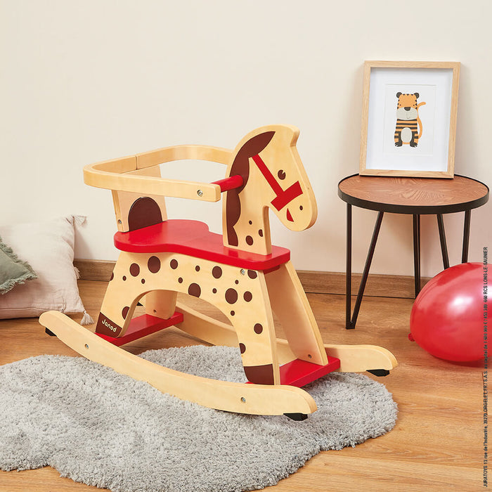 Wooden rocking horse with a red seat, safety rail, and brown spots, sitting on a fluffy grey rug.