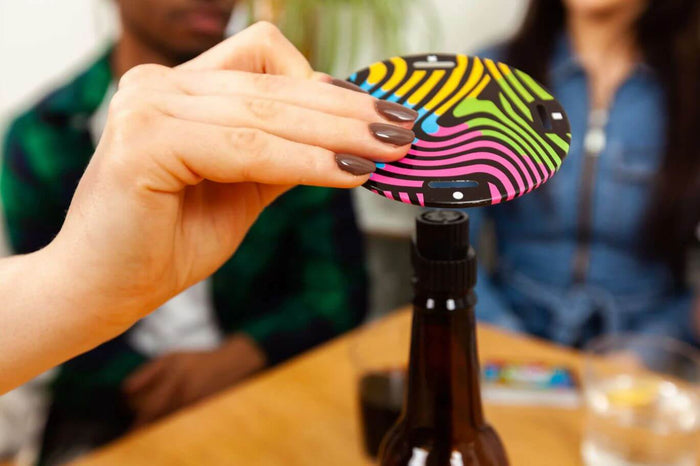 Hand balances a vibrant neon wavy-patterned circular game disc on the top of a dark glass bottle.