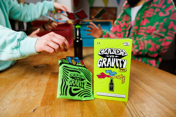 Neon green game box and storage pouch on a wood table with players balancing cards on a bottle in the background.
