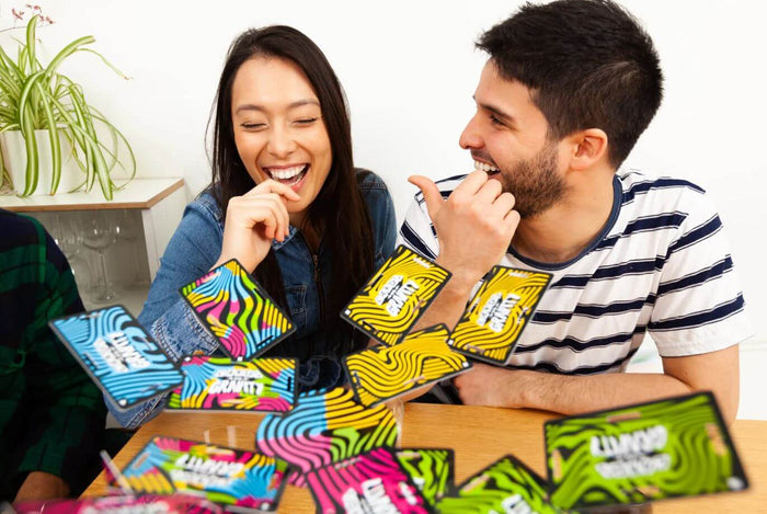 A laughing couple plays with vibrant neon wavy-patterned game cards scattered and tossed over a wooden table.