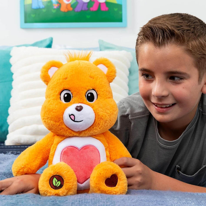 Orange plush bear with a pink heart on its white belly, sitting on a bed next to a smiling boy.