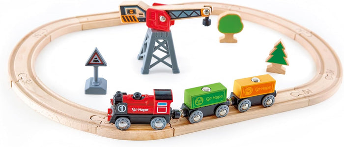 Wooden oval train track with red engine, green and yellow cargo wagons, magnetic crane, traffic sign, and wooden trees.