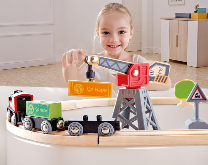 Smiling girl plays with a wooden train set; a grey crane lifts an orange cargo block onto a colorful toy train.