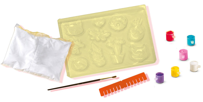 Craft kit with yellow figure mold, white powder bag, paintbrush, orange scraper, and six colorful paint pots.
