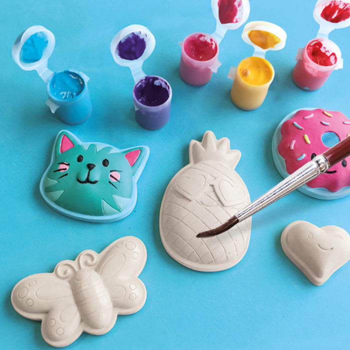 Paint pots and a brush decorating plaster figures of a cat, donut, pineapple, butterfly, and heart on a blue background.