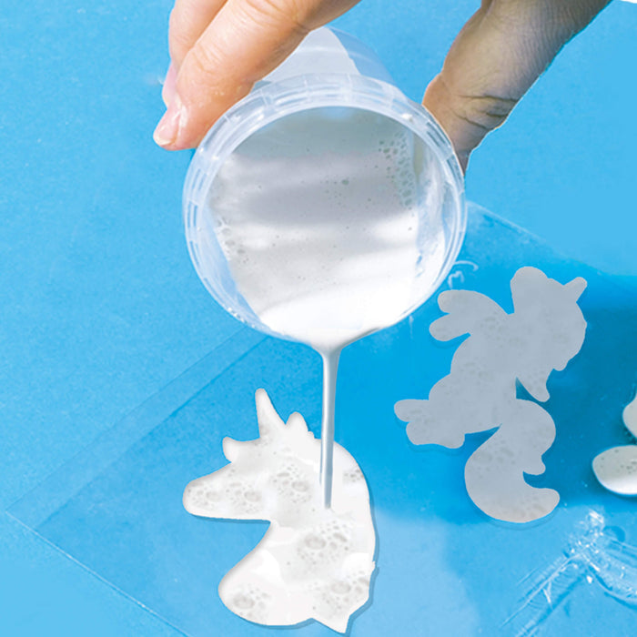 Hand pouring white liquid casting material from a clear cup into a unicorn-shaped mold on a blue background.