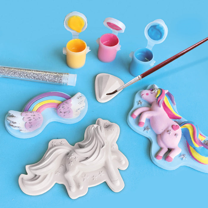 Plaster unicorn and rainbow figures with paint pots, silver glitter, and a paintbrush on a blue background.