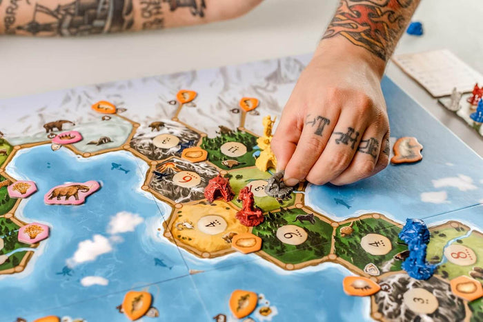 Tattooed hand moving a miniature on a board game with hexagonal tiles, prehistoric animal tokens, and colorful figures.