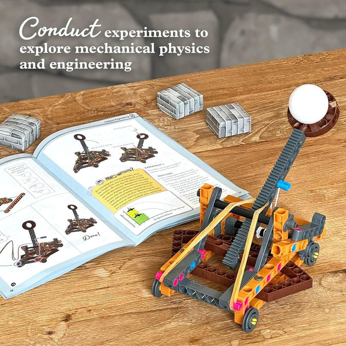 Orange and grey plastic toy catapult loaded with a white ball next to an open instruction manual and toy bricks.