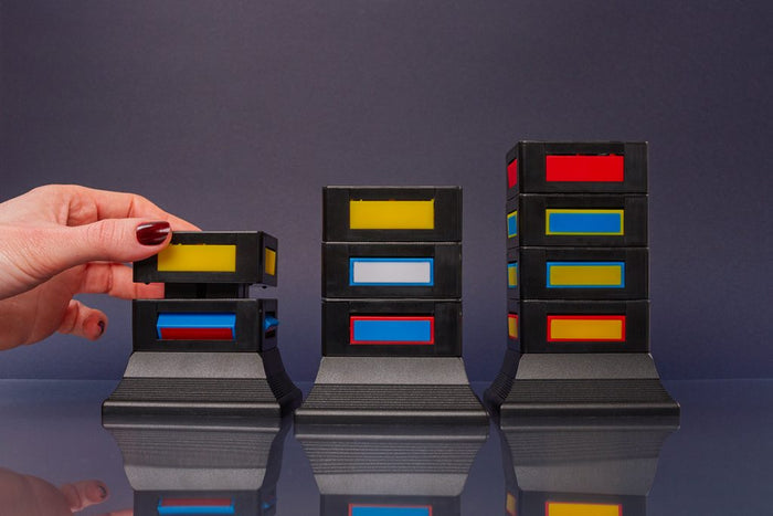 Hand stacking black modular blocks with red, yellow, and blue windows into three towers of varying heights.