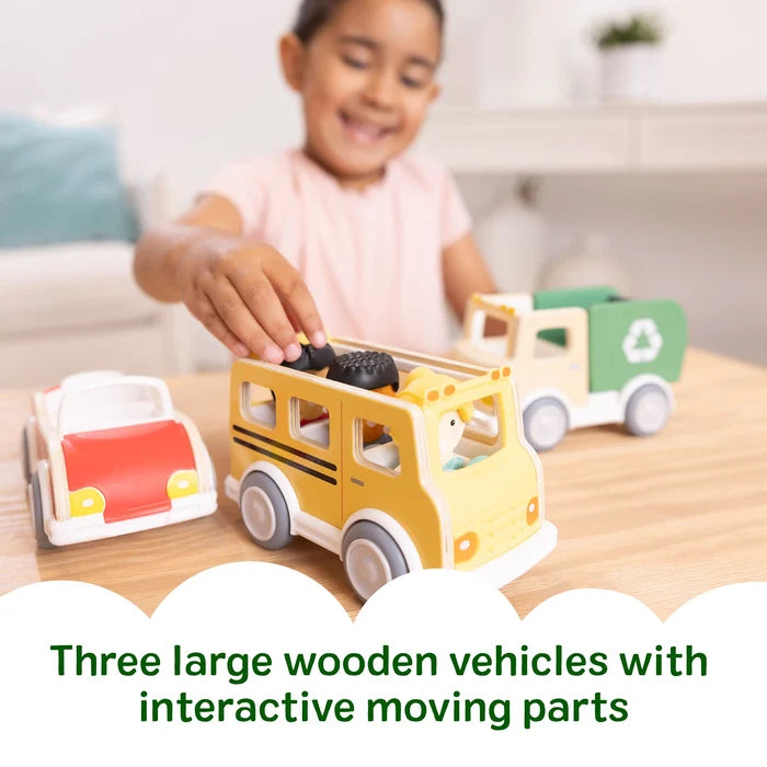 Child plays with a red wooden car, yellow bus with figures, and a green recycling truck on a table.