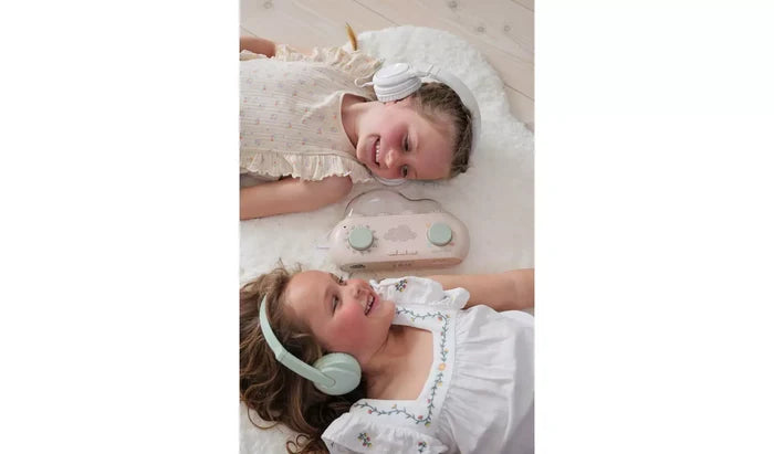 Two girls in headphones lie beside a pink cloud-shaped story box with mint green dials on a white fluffy rug.