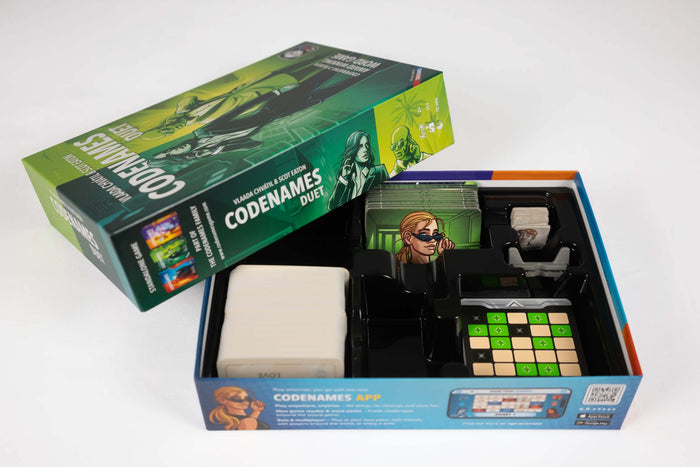 Open green board game box showing cards, character tiles, and a green grid map in a black plastic organizer tray.