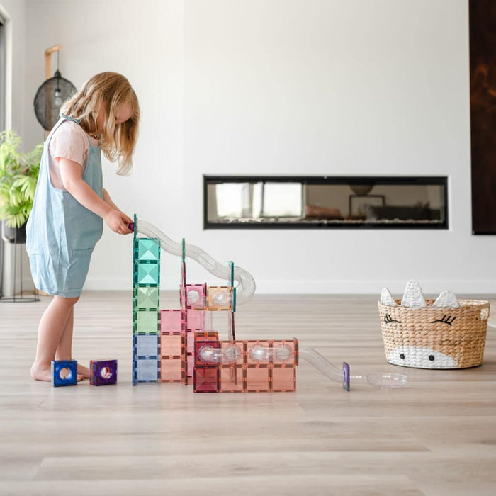 Child building a marble run with pastel magnetic tiles and clear plastic tubes on a wooden floor.