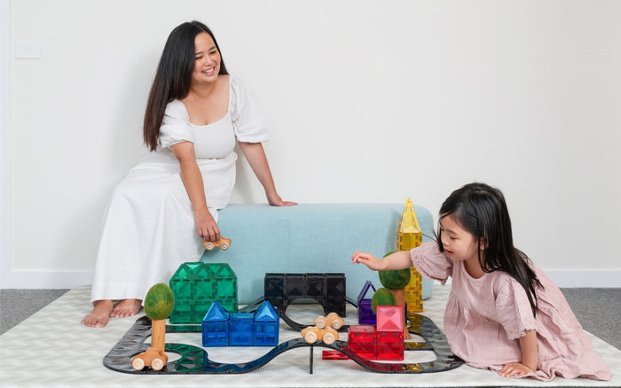 Woman and child build a circuit using black magnetic road tracks and colorful tile houses on a patterned rug.