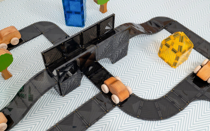Black magnetic tiles formed into a toy road system with a bridge, wooden cars, and trees on a chevron mat.
