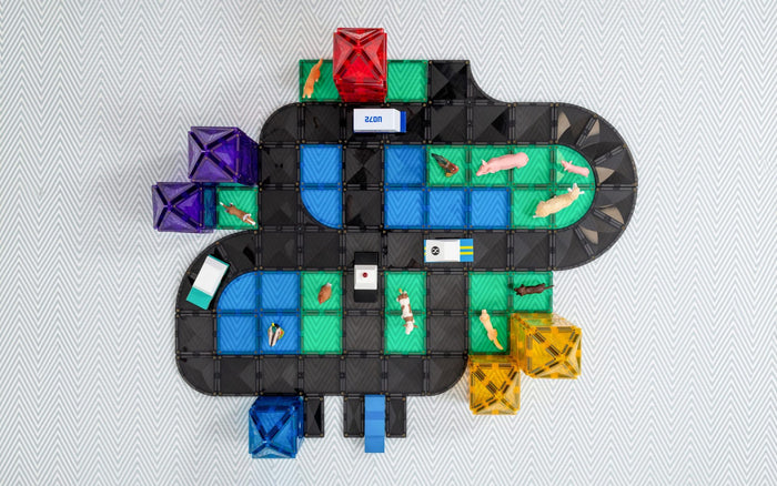 Magnetic tile road layout featuring black tracks, green and blue squares, toy animals, and colorful towers on a rug.