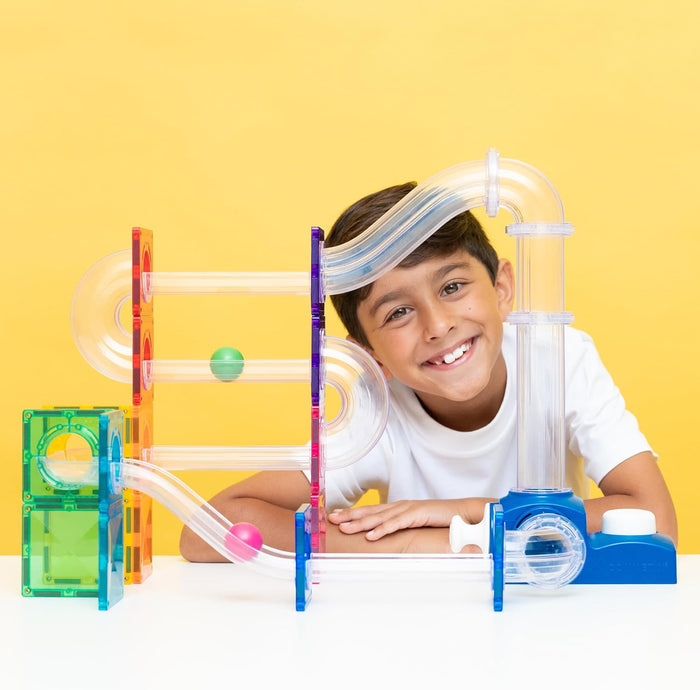Smiling boy with a colorful magnetic tile ball run featuring clear tubes, rolling balls, and a launcher mechanism.
