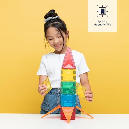 A girl building a rocket with red, yellow, green, and blue light-up magnetic tiles against a yellow background.