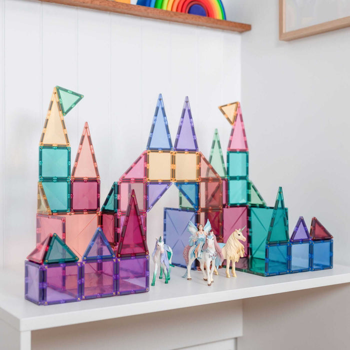 Pastel magnetic tiles built into a castle with towers and arches, featuring toy unicorns and a fairy on a shelf.